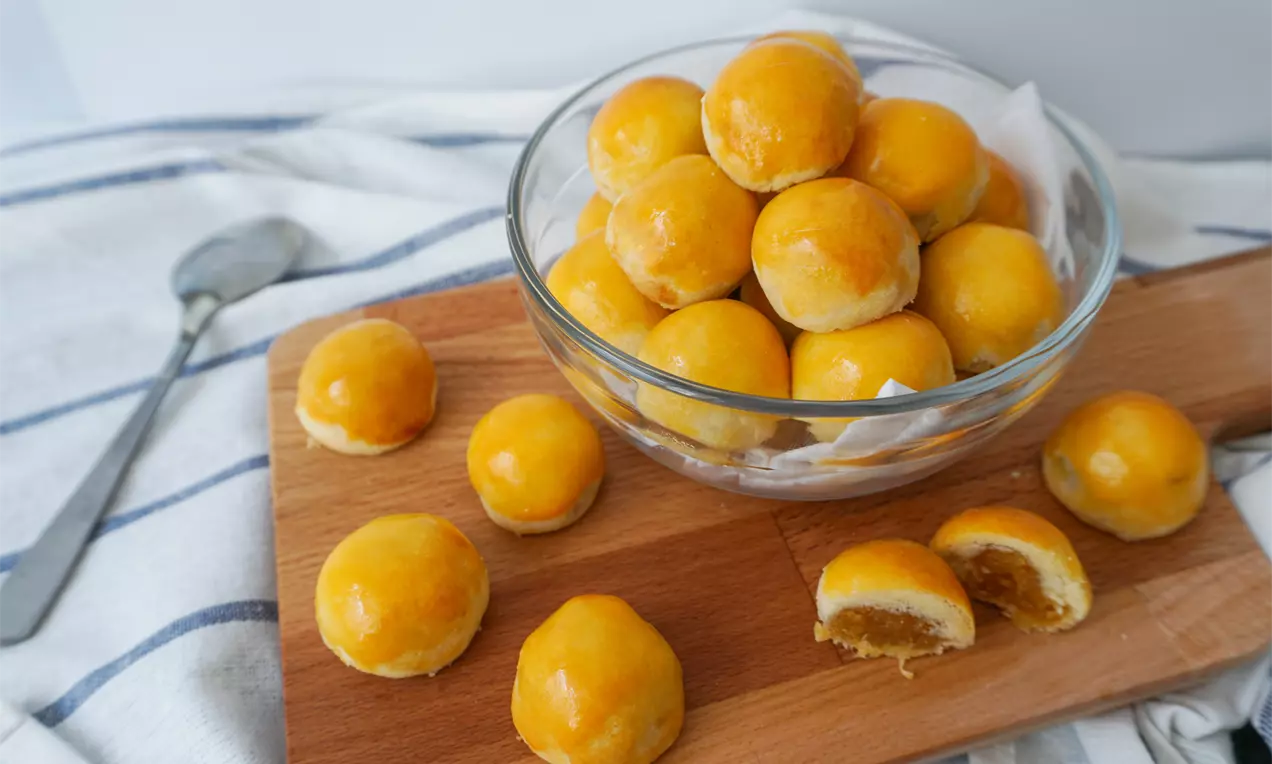 Round golden cookies with a shiny glaze, some with visible fillings, arranged on a wooden board and in a clear glass bowl